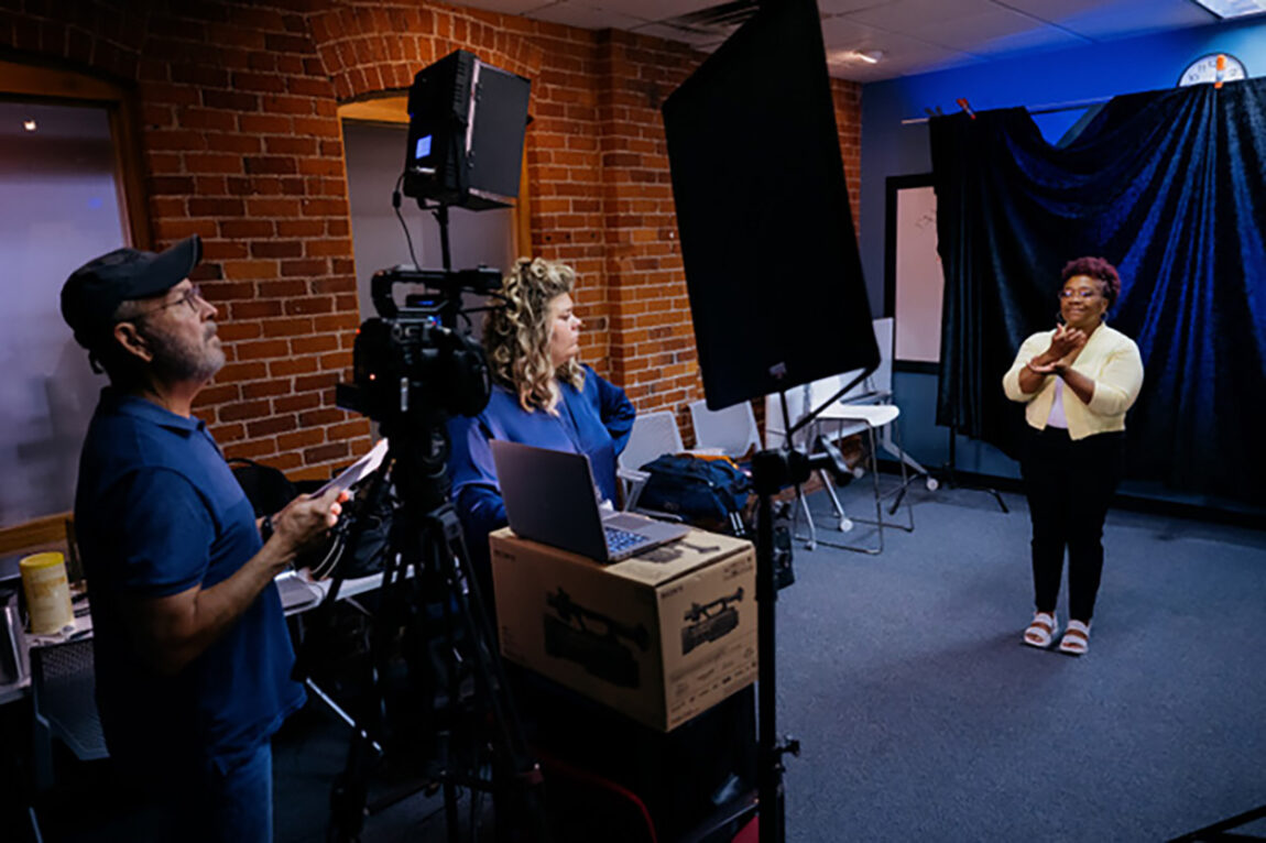Photo of American Sign Language interpreter Stephanie Beatty showing how she provides sign language interpretation for President Santa J. Ono’s June video message, as ASL interpreter Casie Watson toggles through digital cue cards and Michigan Media executive producer Bob Berg looks on.