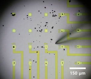 A rectangular array of 20 pale-green squares, which are the array’s microelectrodes. Some arrays have slightly darker wires extending to the edge of the image. Each microelectrode with a wire has a black dot (battery particle) on it. The black dots also speckle the image, and a blurred microneedle near the center of the array has been used to push the particles onto the electrodes.