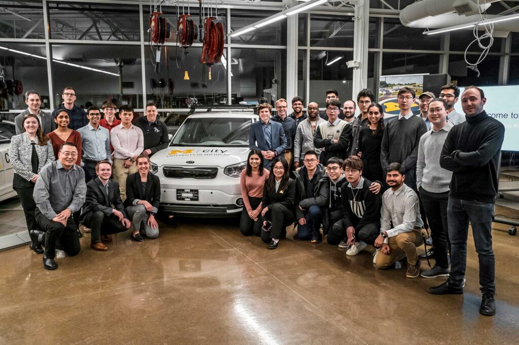 About 30 people surround a white car with Mcity on the hood, in a brightly lit building of steel and glass.