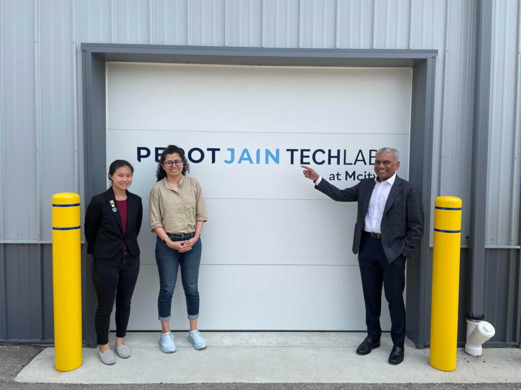 wo women and a man stand in front a building sided in gray corrugated metal, with the man pointing at the "PEROTJAIN TECHLAB at Mcity" logo on the white sectional garage door.