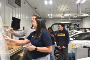 A man with a marker smiles at the whiteboard while a woman points to something, also smiling. Behind them, a woman and a man look at something hidden behind the woman at the whiteboard. In the background is a white car with Mcity on the side. The building has white corrugated metal walls, tall garage doors and bright strip lights.