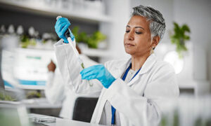 Woman works in lab dressed in white coat and gloves holding testtube. She has olive skin and short gray hair.