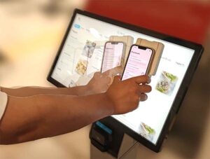 A touchscreen monitor displays several food items on a menu. The monitor's screen is backlit and the frame is black. Someone is holding a smartphone connected to BrushLens against the screen while moving it from left to right. BrushLens appears as an off-white case around the edges of the phone.