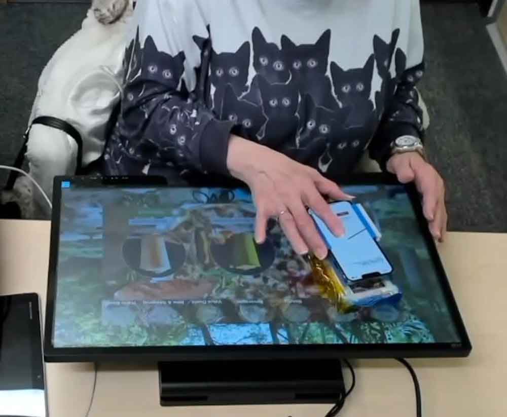 A person is sitting at a tan-colored desk. A black-bordered screen lays flat on the desk and displays a green menu with several images of drinks. Several black cords are connected to the screen and run off the desk. The person is moving a phone inside a BrushLens phone case across the screen. A red arrow is point to her menu choice in the top right corner of the screen, from the viewer's perspective.