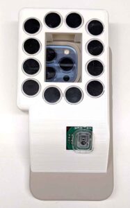 The picture shows the underside of BrushLens, a white phone case. A window in the center of the case reveals a smartphone camera. Thirteen black circles, which are the clickers, surround the window in a circle.