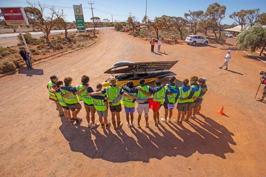 Fourteen members of the U-M race crew stand together in a line while linking arms. Their backs are facing the viewer. Each team member is wearing a neon green vest with reflective safety markings. In front of the crew is the solar car, Astrum, which has a yellow body. The top panel of the car is opened so that the solar panels are facing the sun. With the panel opened, the inside of the car's black cockpit is visible.