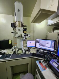 The transmission electron microscope is a long, tan device standing upright on a black desk. Several knobs and attachments protrude from the instrument, and a collection of wires connect the device to a computer panel on the right-hand side of the microscope.
