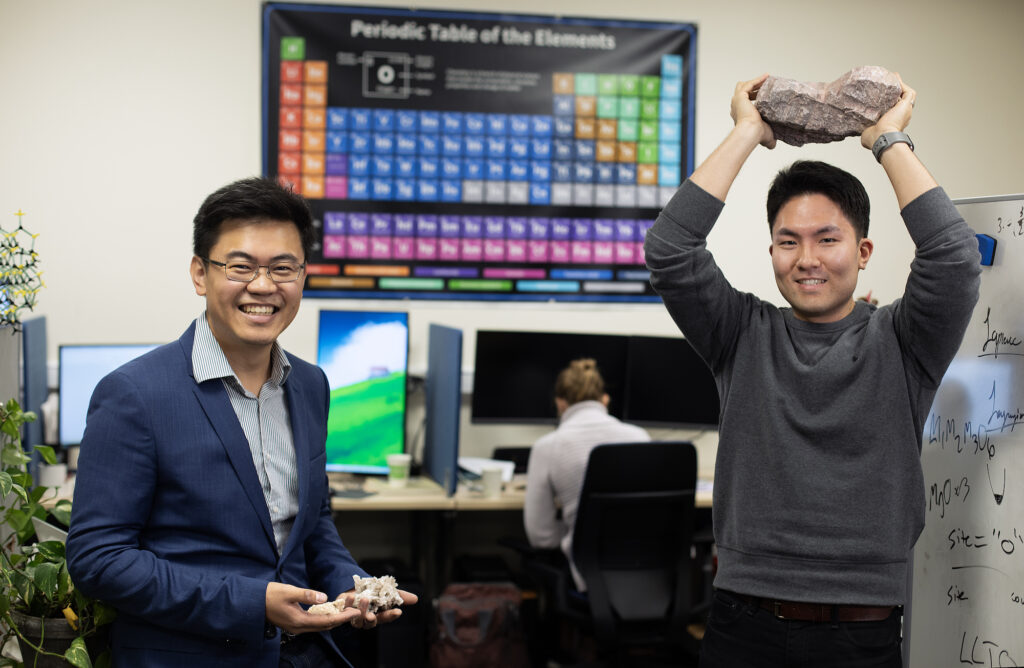 Two men holding rocks stand in a computer laboratory. The man on the left is wearing black-rimmed glasses and a navy blue blazer over a white shirt with fine black stripes. He also holds two palm-sized, light-pink rocks in each hand. The man on the right is holding a larger, light-pink rock over his head and is wearing a gray sweater.
