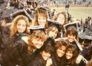 Group of females with hairdos circa 1989 prepare for commencement in the big house. They are dressed in caps and gowns. Field in the background.