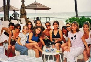 Circle of women at a pool with ocean and umbrella in the background.
