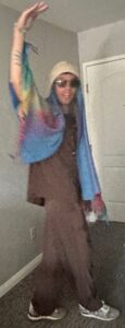 A guy in sunglasses is draped in a tie-dyed cape and wacky cap.