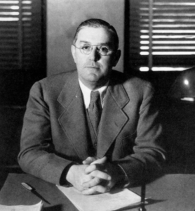 Middle-aged caucasian man, circa 1945, sitting at a desk, hands folded, wearing glasses with hair slicked back. In a suit and tie. Black & white.
