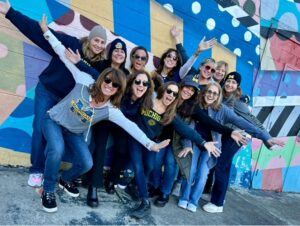 Female Michigan Grads in U-M gear pose gleefully in front of a mural in Ann Arbor.