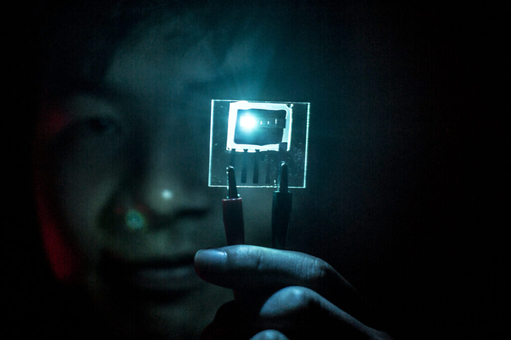 A researcher uses forceps to hold up a small chip in a dark room. The chip contains a blue PHOLED, which lights up the surrounding area. The lighting makes the researcher's hands and face appear a blue-green color.
