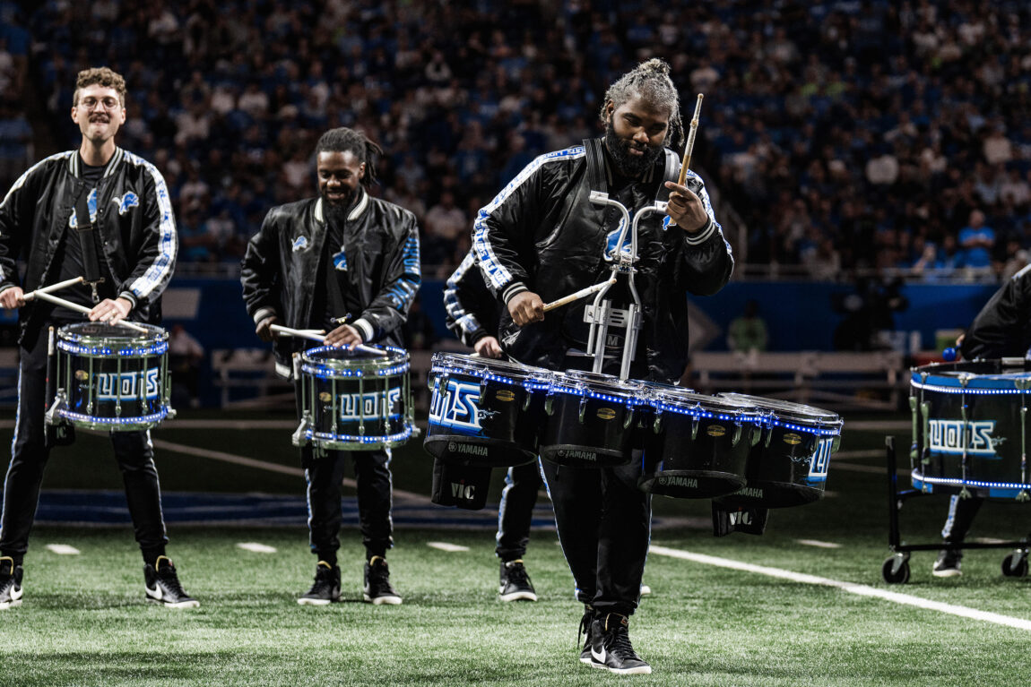 A photo of three men playing drums inside Ford Field during a Detroit Lions football game.
