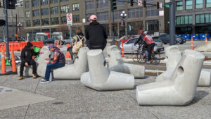 People sit on an outdoor sculpture comprising several interlocking pieces that can be used to support seawall infrastructure in Seattle. Urban scene with oddly shaped sculpture.