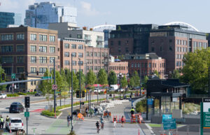 A longshot of a Seattle street that features the sculpture of Buster Simpson. Industrial brick buildings, a crosswalk, several people, and cars.