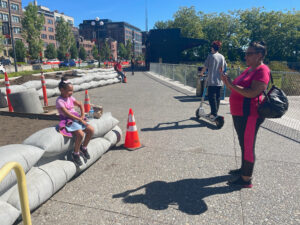 A woman photographs a child on a bright sunny day. The child is sitting on an outdoor sculpture that appears to be sandbags.