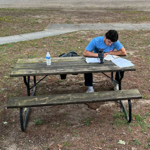 Young man with head bowed over books sits at a picnic table outside.