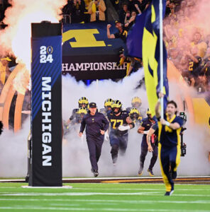 Athletes emerge from the tunnel onto the football field, surrounded by smoke.