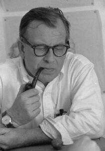 Black & white image of man in white shirt and horn rim glasses, sleeves rolled up, smoking a pipe.