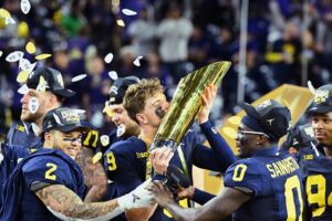 Michigan football players surround national championship trophy as the one in the center gives it a kiss. The trophy is tall, cylindrical, and a golden color.