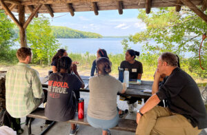 Group of eight people converse at a picnic table under a pavilion on a sunny day with a beautiful lake in the background.