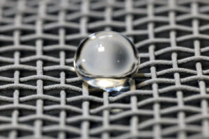 A water droplet rests on gray mesh, reflective and translucent like a cloudy glass marble.