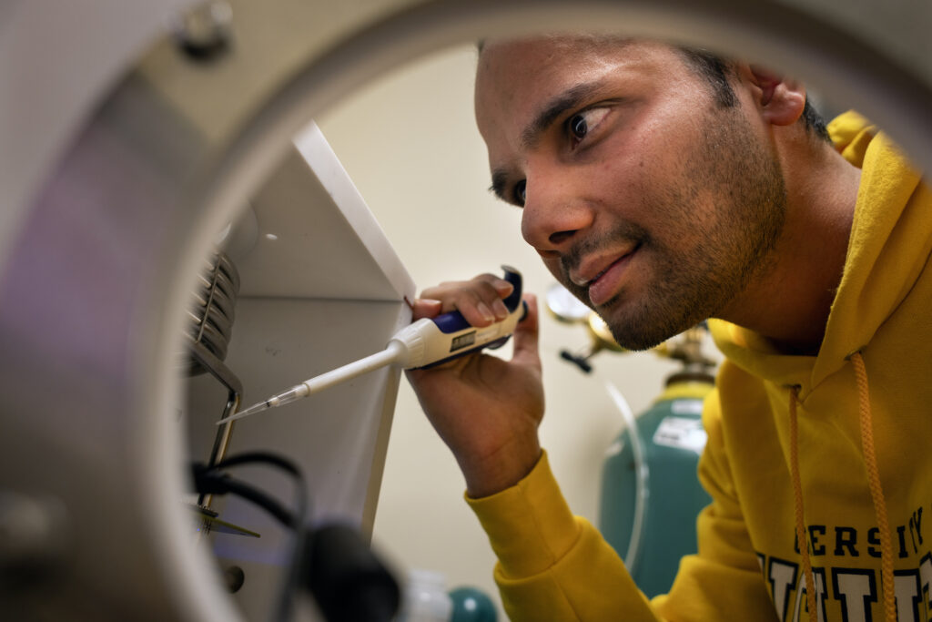 Sudeep Sharma peers into a white cabinet. He is holding a micropipette, which has a plunger instead of a bulb. The transparent, plastic tip of the micropipette is aimed into the box.