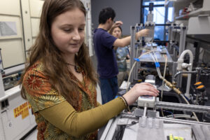 Olsen rests her hand on a force gauge, which is connected to a steel block by a retractable metal rod. The block is aligned with the end of an ice block, which rests on a metal surface. The rod is pushing the blocky end of the hammer toward the ice block.