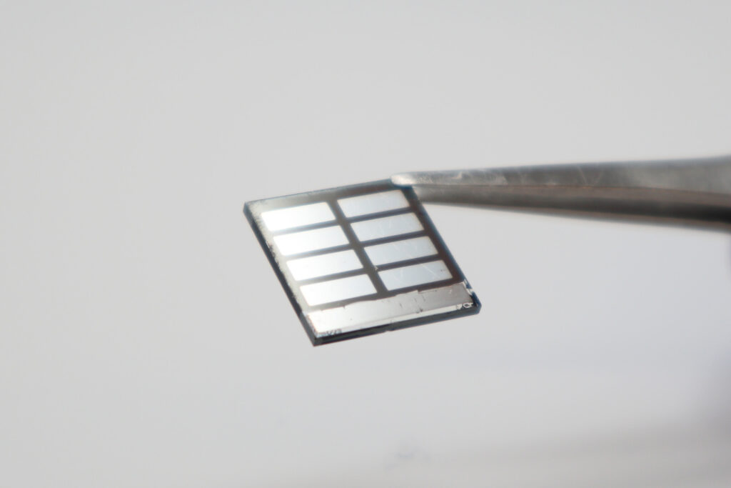 A pair of forceps hold a perovskite solar cell, which looks like a thin, square panel. The cell's electrodes look like silver rectangles on the cell's surface, and the surrounding black surface is the perovskite film.