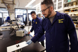 Pena-Francesch and Zhang stand at a black lab bench. On the benchtop, a tan electrical box about the size of a loaf of bread is connected to a coil via red and black wires. The coil resembles an upside-down, metal cup. The soft robot sits atop the coil, resembling two black band-aids that cross at their centers. Pena-Francesch holds a white sheet of paper between the soft robot and the coil, to provide contrast with the black table top.