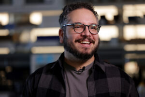 A portrait of Abdon Pena-Francesch standing in front of a set of shelves in his laboratory. He wears a beard, flannel shirt, black-rimmed glasses, and a big, bright smile.