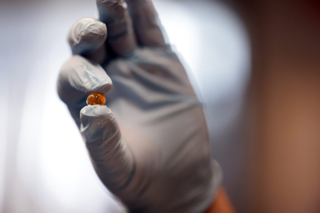 A gloved hand holds an orange capsule in between thumb and index finger.