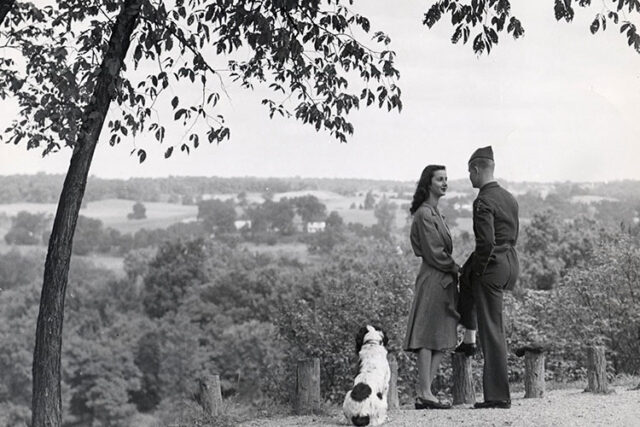 Photo of a dog, woman and uniformed soldier overlooking trees and farmland