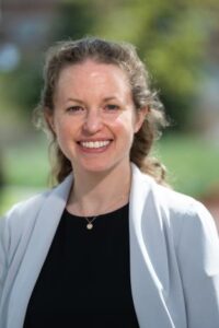 Headshot of smiling caucasian woman with pulled-back hair standing outside in front of greenery.