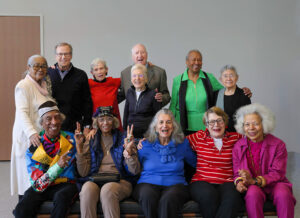 A group photo of racially diverse elderly people dressed in bright colors, smiling, and flashing peace signs.