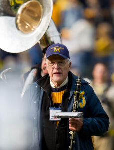 Elderly man wearing a blue cap with maize block M that reads Alumni Band holds a wind instrument and prepares to perform during Homecoming Weekend, 2011.