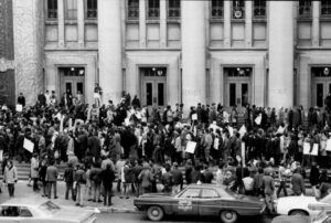 Crowd of protesters gather on the steps of Hill Auditorium carrying signs. Vintage autos on the street, circa 1970.