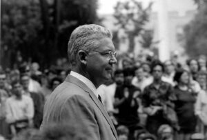 Profile of caucasian man with gray hair and glasses in foreground with student protesters in the background