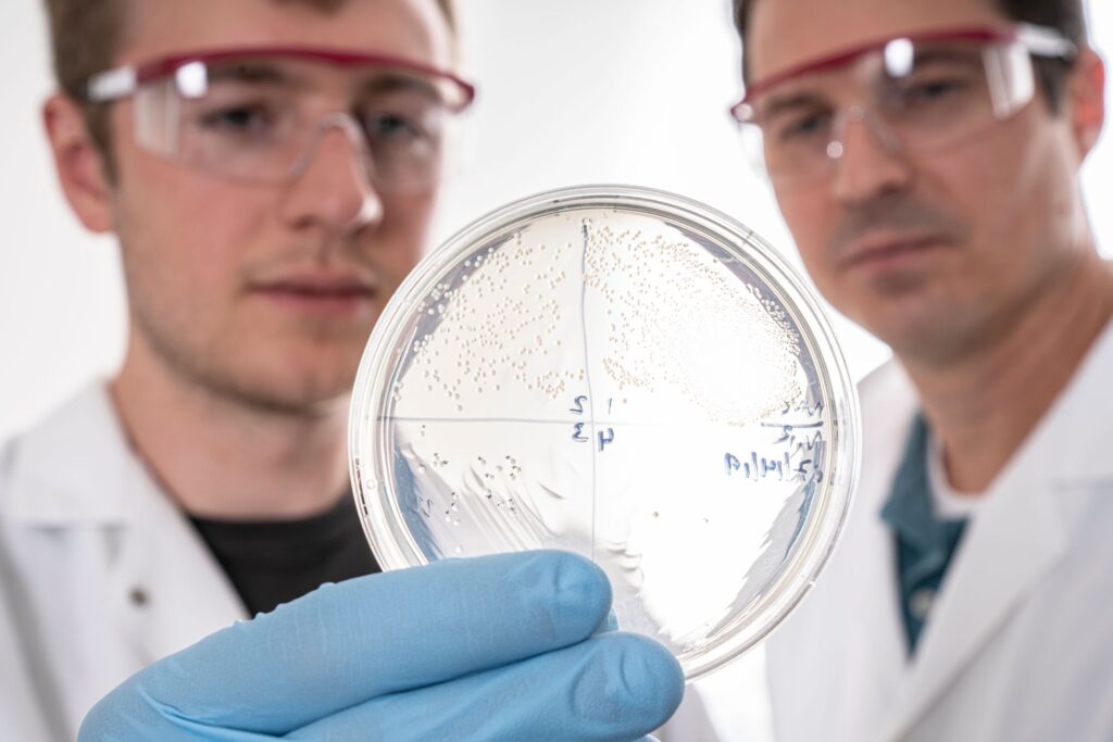 A round, clear plastic dish with a clear, speckled film inside is in the foreground. Two men in safety glasses are out of focus in the background.