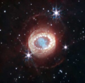 On a black backdrop, dim or distant stars glow white, with three nearer or brighter stars producing hexagonal diffraction patterns like rainbow snowflakes. In the middle of the frame, the supernova remnant resembles a reptilian eye, with a teal cloud in the center ringed by bright white dots that are the clumps of hydrogen lit up by the shockwave from the supernova. A pale haze surrounds the ring of dots, with other less distinct dots inside. A near-infrared image of the remnant left behind by supernova 1987A, taken by the James Webb Space Telescope. The hydrogen clumps known as the "string of pearls" appear as a ring of white dots around the teal center of the stellar remnant, still shining brightly due to the energy imparted by the supernova shockwave. The number of clumps is consistent with the Crow instability having caused them to form. Image credit: NASA, ESA, CSA, M. Matsuura (Cardiff University), R. Arendt (NASA’s Goddard Spaceflight Center & University of Maryland, Baltimore County), C. Fransson (Stockholm University), J. Larsson (KTH Royal Institute of Technology), A. Pagan (STScI)