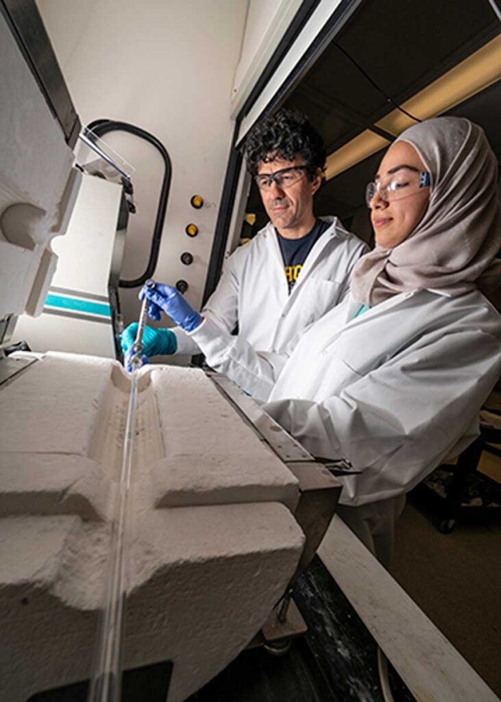Two scientists stand in front of a furnace that is sitting on a lab bench. The furnace looks like a metal box and the top half is opened by a hinged door. Inside the furnace is white ceramic material. The reactor, which resembles a glass tube, sits inside a channel carved into the ceramic.