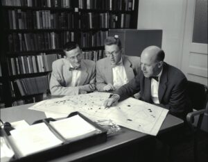 Three men examine a map. They are all wearing bow-ties, circa 1956. Black & white.