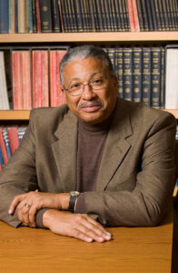 Smiling African American scholar sits at table with arms folded, thoughtful.