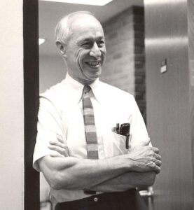 Smiling man, older, arms crossed, striped tie. Black & white.