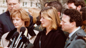 Two caucasian women amid a crowd speak into a cluster of microphones.