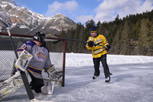 Hockey player scores goal, dressed in maize. Outside on a rink surrounded by pines and mountains.