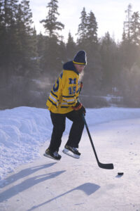 Hockey player catches air. Dressed in maize jersey on an outside rink rimmed by pines.