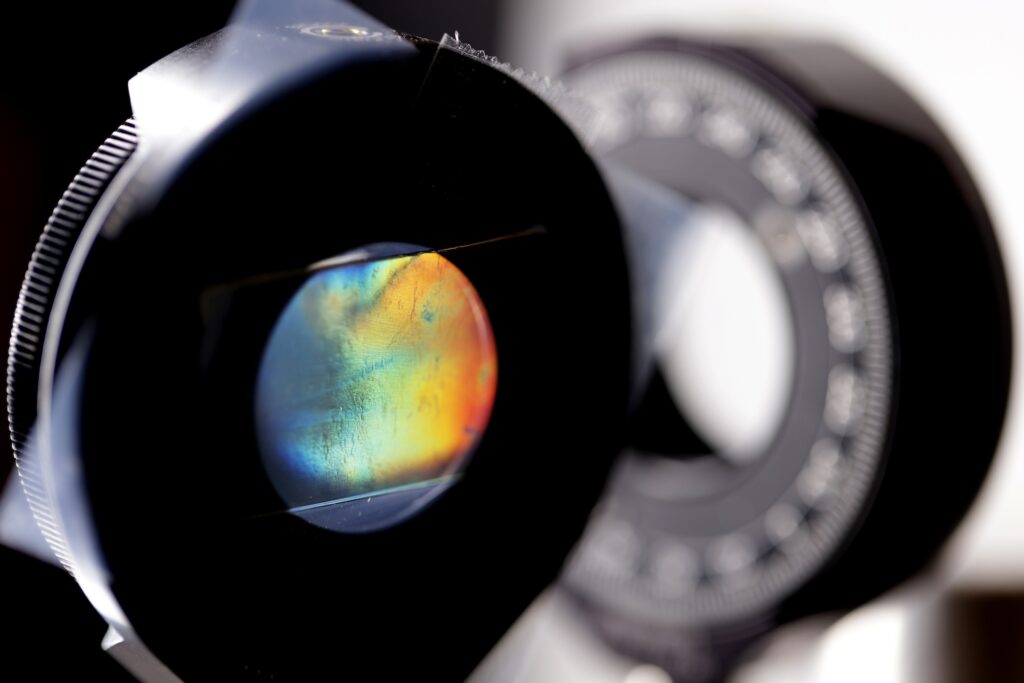 A thin sheet of material is sandwiched between two black, donut-shaped clamps. Through the hole, the sheet's shimmering color shifts from blue to red.